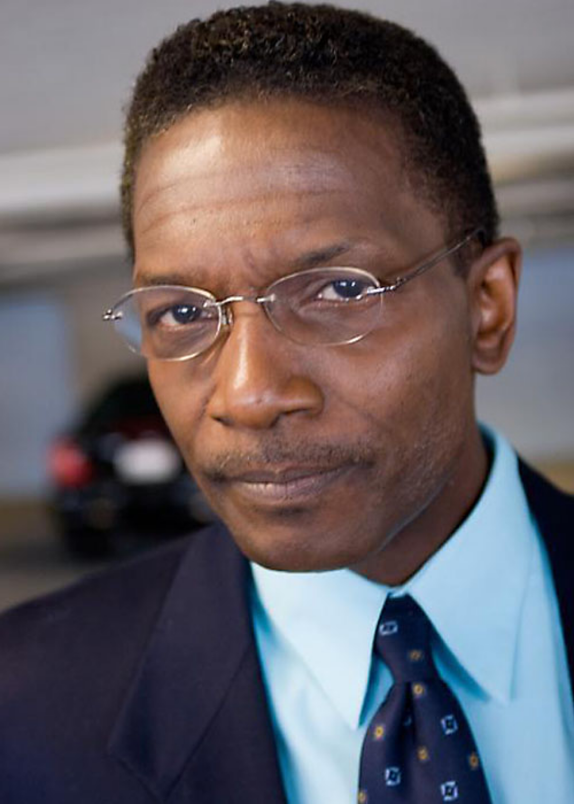 Sedly Bloomfield in a dark suit, blue shirt, tie, and glasses